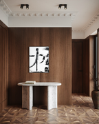 A framed black and white abstract calligraphic wall art print hangs on a dark wood-paneled wall above a white stone console table in a modern interior hallway with patterned wood floors.